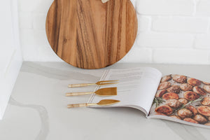 Wood charcuterie board with personalization available, Appetizer and charcuterie cookbook, idea book for charcuterie board, brass and rattan wrapped cheese utensils, white countertop, Mercy Forest Co., custom gifting for realtors, on-demand ready-to-ship gifts, packaged gift box