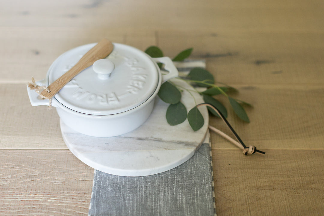 White brie baker with wooden knife, white and grey marble trivet, and linen tea towel on wooden background. Realtor gifts. Housewarming gifts. Mercy Forest Co.