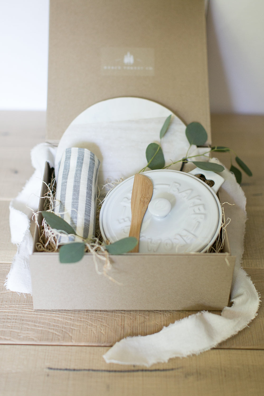 White lidded brie baker with wooden spoon, white and grey marble trivet, and a striped linen tea towel neatly arranged in brown box. Realtor gifts. Housewarming gifts. Mercy Forest Co.