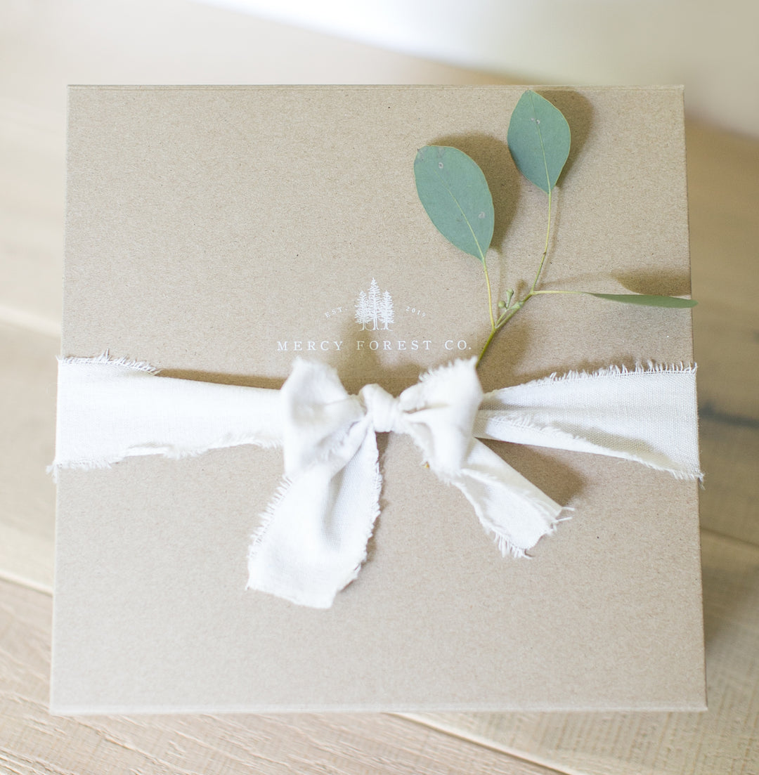 Kraft paper box with Mercy Forest Logo, a white linen ribbon tied around it, with a sprig of eucalyptus on wooden background. Realtor gifts. Custom gifts. Housewarming gifts. Mercy Forest Co.
