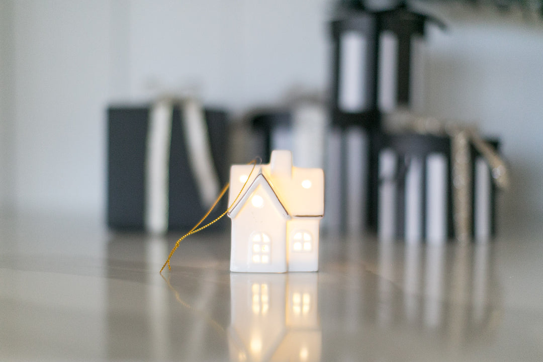 Decorative house-shaped ceramic ornament with led light on a reflective surface with blurred gift boxes in the background