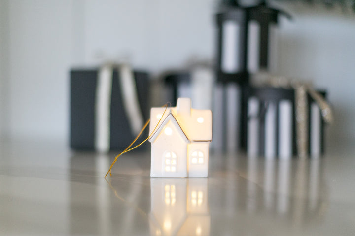 Decorative house-shaped ceramic ornament with led light on a reflective surface with blurred gift boxes in the background