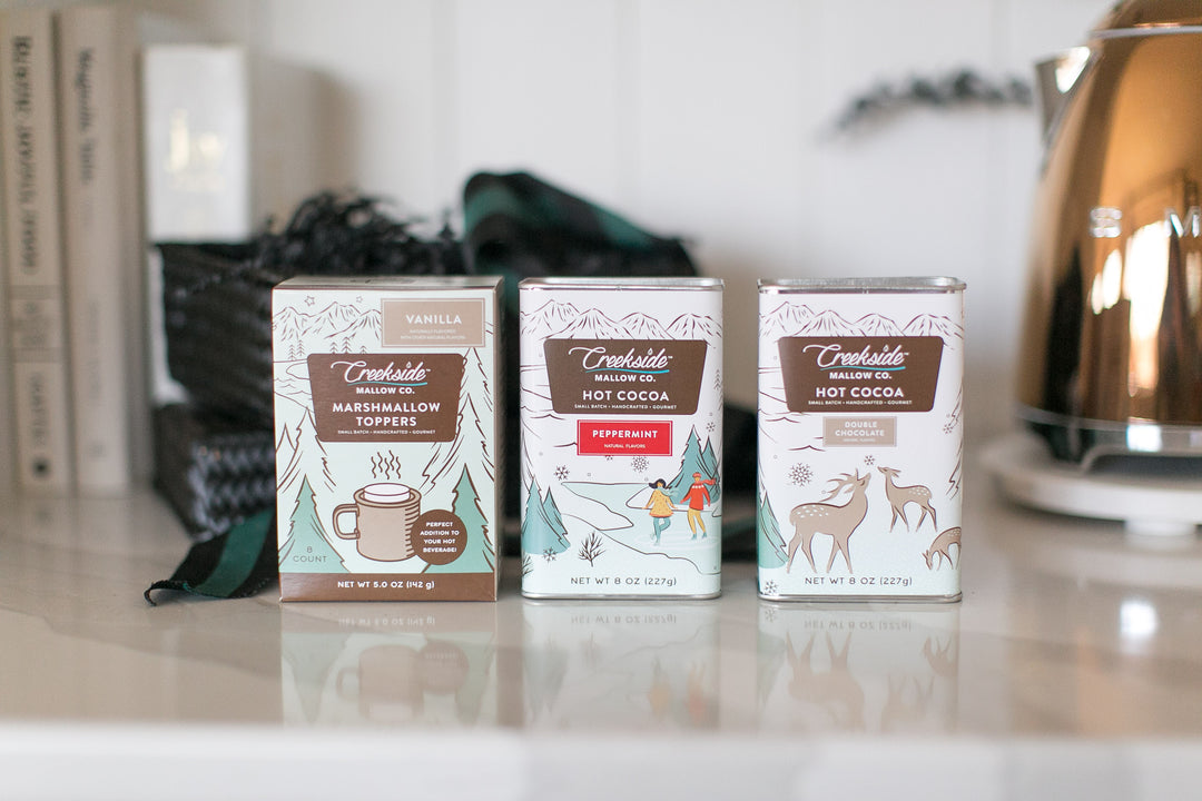 Three boxes of Creekside Mallow Co. Hot Cocoa on a kitchen counter with a kettle in the background.