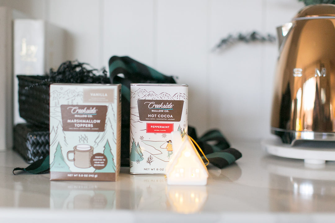 Two boxes of creekside mallow co. hot cocoa on a kitchen counter with a kettle in the background and ceramic LED ornament in front