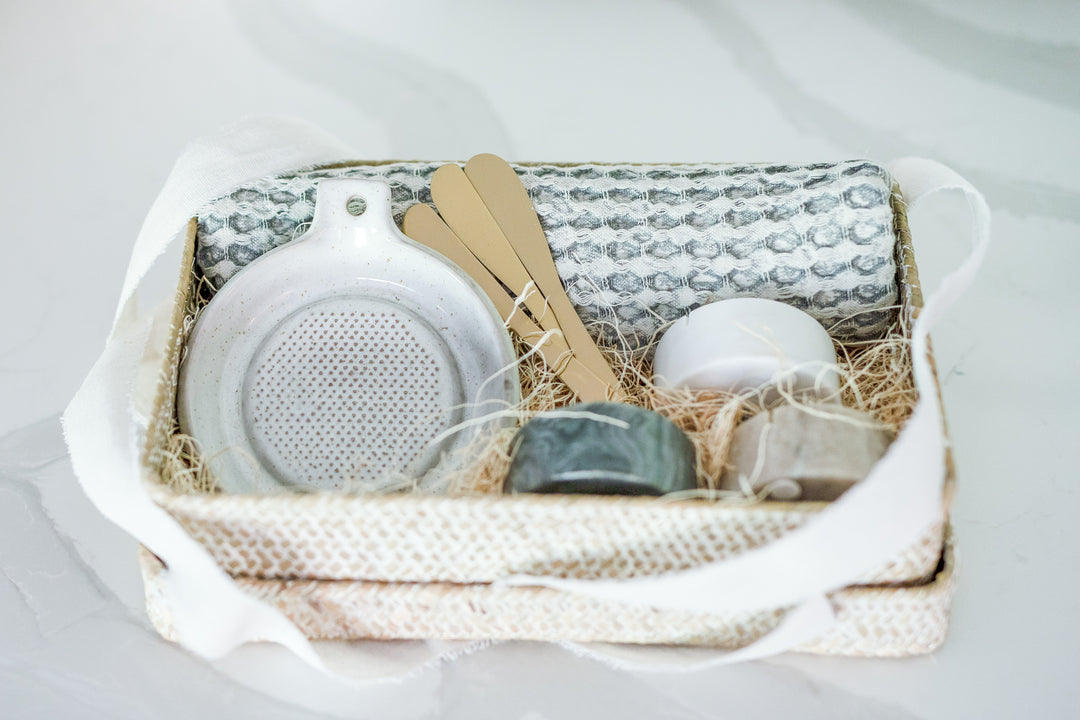 Mercy Forest Co. gift box, ready to ship, displayed in a woven seagrass box with a lid. Inside are three elegant marble dishes in shades of white, tan, and charcoal, each paired with a brass spreading knife. Also included are a stoneware grater and a soft, cotton waffle weave kitchen tea towel, creating a refined and thoughtful gifting ensemble.