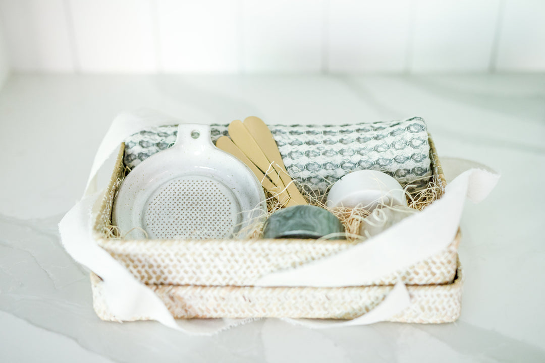 Mercy Forest Co. gift box in a woven seagrass container with a lid, arranged and ready to ship. Inside, the gift includes three marble dishes in shades of white, tan, and charcoal with matching brass spreading knives, a stoneware grater, and a cotton waffle weave kitchen tea towel. The items are thoughtfully arranged to highlight the quality and style of each piece.