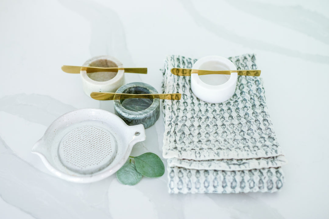 Mercy Forest Co. gift items displayed on a marble countertop, featuring a set of three marble dishes in white, tan, and charcoal, each with a brass spreading knife. Accompanying the dishes are a stoneware grater and a soft, cotton waffle weave kitchen tea towel, arranged to showcase the elegant and cohesive design of the gift set.