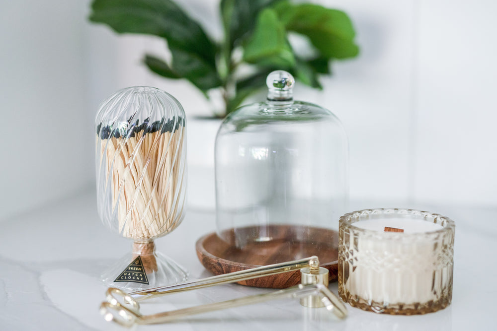 A beautifully arranged gift set featuring a round wood dish with a glass cloche, holding a 7oz Oak & Ember candle in a reusable glass vessel. The candle has a signature scent of vanilla and sandalwood. Beside it, there's a brass wick trimmer, brass candle snuffer, and a decorative jar containing a set of 100 matchsticks. All items are placed in a sturdy round lidded box adorned with stuffing and a signature Mercy Forest Co. ribbon, showcasing a reusable woven paper box, perfect for storage.