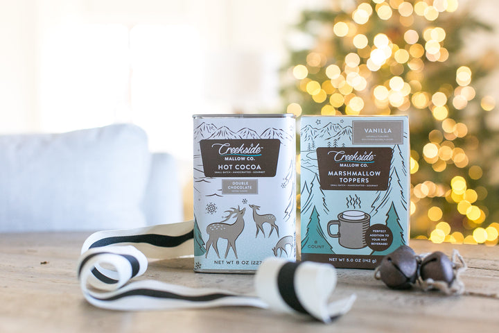 Two boxes of Creekside Mallow Co. hot cocoa and marshmallow toppers on a wooden surface with a blurred festive background.