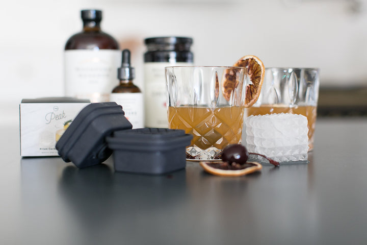 Glass of whiskey with ice cubes, bottles, and a box on a reflective surface.