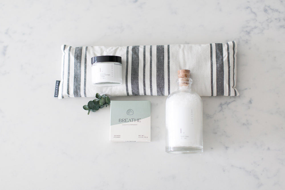 A grey and white eye mask, eucalyptus shower steamer, bottle of bath salts, and lavender body cream on white and grey marble with sprig of eucalyptus. Relaxation gifts. Mercy Forest Co. Gifting ideas. 