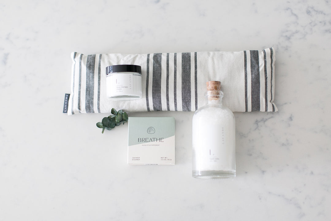 A grey and white eye mask, eucalyptus shower steamer, bottle of bath salts, and lavender body cream on white and grey marble with sprig of eucalyptus. Relaxation gifts. Mercy Forest Co. Gifting ideas. 