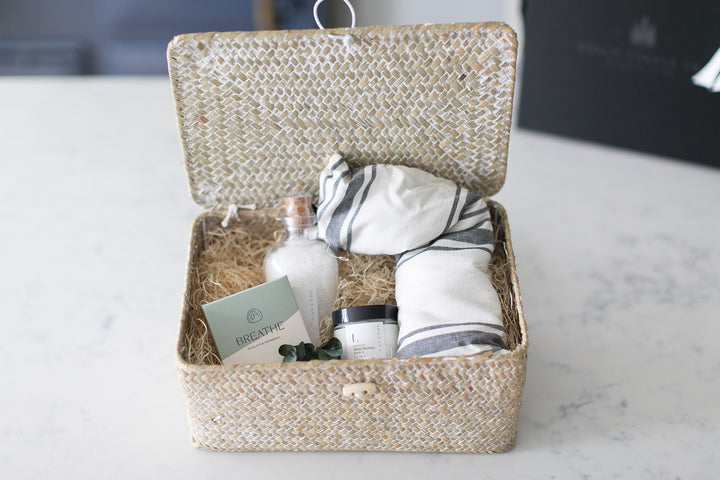Hinge top gift basket with grey and white striped eye mask, lavender bath salts, a shower steamer, and lavender body cream on a white and grey marble table. Relaxation gifts. Pampering gifts. Mercy Forest Co.