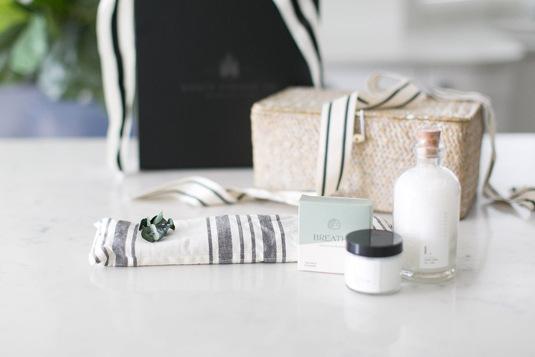 A wicker basket containing salt soak bottle, a jar of lavender body cream, a shower steamer in eucalyptus, a lavender eye pillow, all neatly arranged on a white surface with a black and white striped cloth  ribbon on top. Relaxation gifts. Pampering. Mercy Forest Co. 