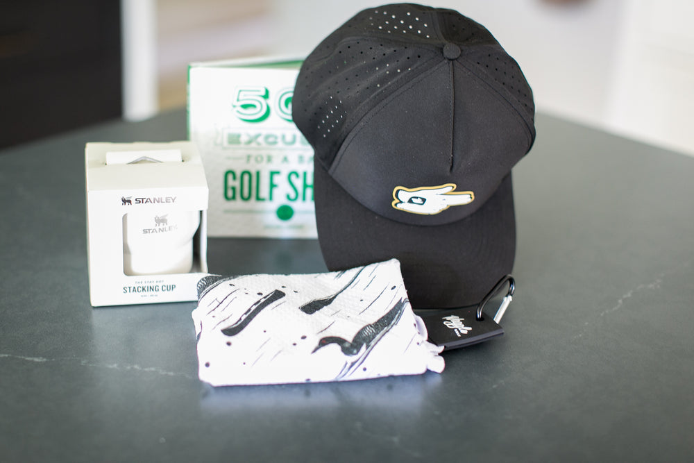A black perforated Waggle golf shooter baseball cap with a gold and white logo, displayed next to a waggle white and black loon patterned golf ball towel, a white Stanley stacking cup in its packaging, and a green and white golf shot book on a dark gray tabletop.