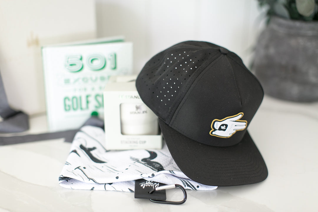 A black waggle shooter baseball cap with a gold and white logo, displayed on a white surface next to a patterned towel golf ball towel, a white Stanley stack and go mug in its packaging, and a golf book in the background.