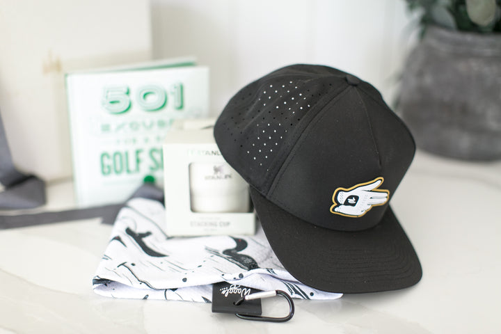 A black waggle shooter baseball cap with a gold and white logo, displayed on a white surface next to a patterned towel golf ball towel, a white Stanley stack and go mug in its packaging, and a golf book in the background.