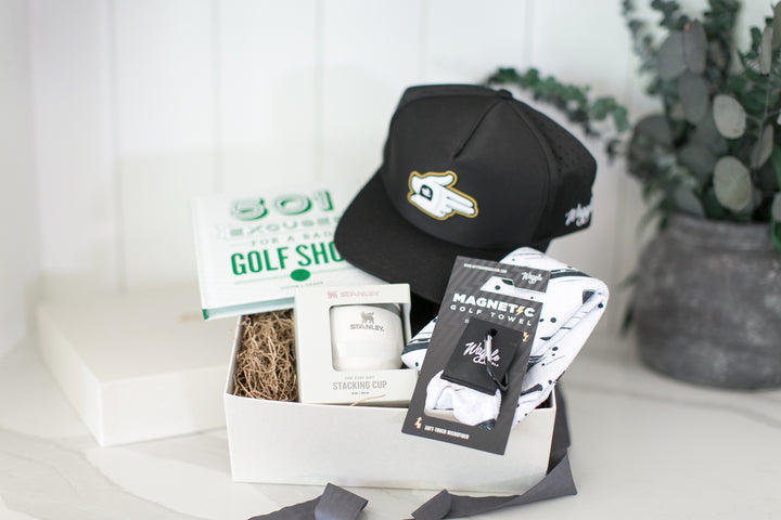A curated gift box set featuring a black baseball cap with a gold and white logo, a white tee box with a white tees cup, a golf shop card, and a packaged pair of golf socks, arranged in a white box with decorative twine on a light surface.