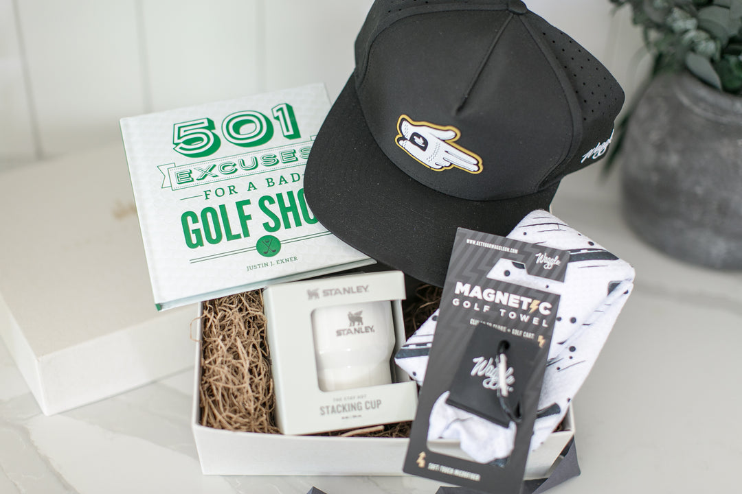 A curated golf gift box set on a light marble surface. The set includes a black Waggle baseball cap with a white and gold golf ball shooter logo, a white book titled '501 Excuses for a Bad Golf Shot', a white Stanley stacking cup in a box, a magnetic black and white golf ball towel, and a white gift box.