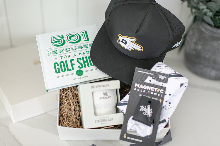 A curated golf gift box set on a light marble surface. The set includes a black Waggle baseball cap with a white and gold golf ball shooter logo, a white book titled '501 Excuses for a Bad Golf Shot', a white Stanley stacking cup in a box, a magnetic black and white golf ball towel, and a white gift box.