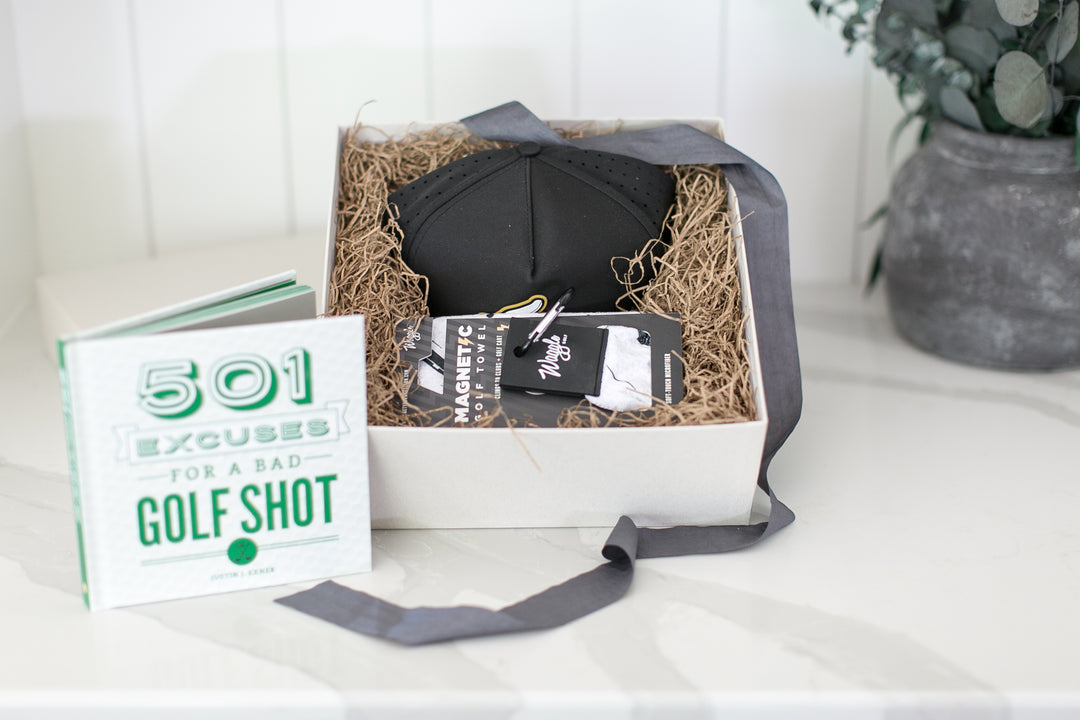 A curated golf gift box set on a white marble countertop. The set includes a white book titled '501 Excuses for a Bad Golf Shot', a black golf hat, a black and white patterned golf ball bag, and a gray ribbon, all nestled in brown paper shred inside a white box.