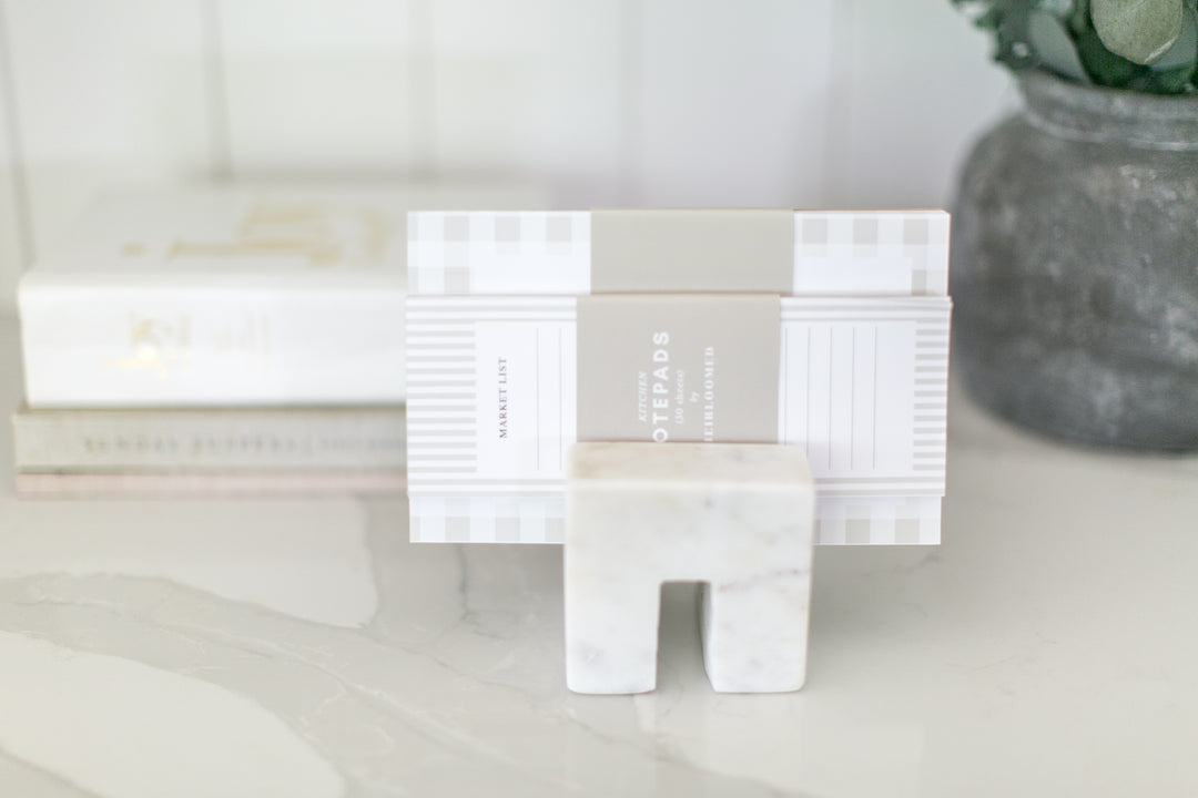 A white and gray geometric desk organizer stand holding a notebook, set on a marble surface with a blurred gray pot in the background.