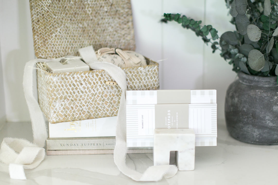 A styled product display featuring a woven gold and white basket with a white ribbon, stacked books, a white and gray patterned box, a matching small marble block, and a white ribbon on a light marble surface next to a potted eucalyptus plant.