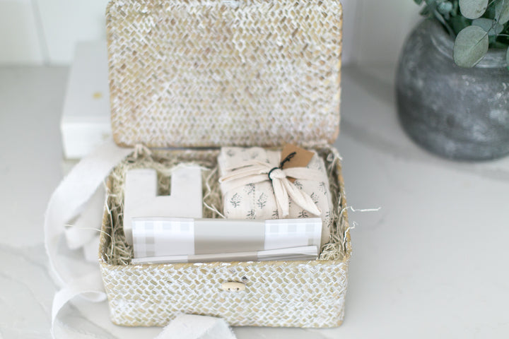 A decorative gift box with a textured beige and white pattern, shown open on a light surface. Inside, a wrapped gift tied with a ribbon and a small tag are nestled in shredded paper filler, alongside white decorative blocks and a folded paper item.