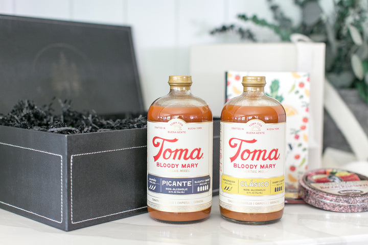 Two bottles of Toma Bloody Mary seasoning mix on a white surface, next to a black gift box with shredded paper.