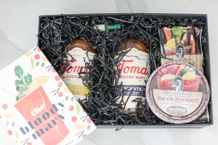 A curated gift box set containing two bottles of Toma Bloody Mary Mixer and a bottle of Demitris Bacon Rim Shot, nestled in black shredded paper inside a black gift box, with a printed 'the bloody mary' card on the left.