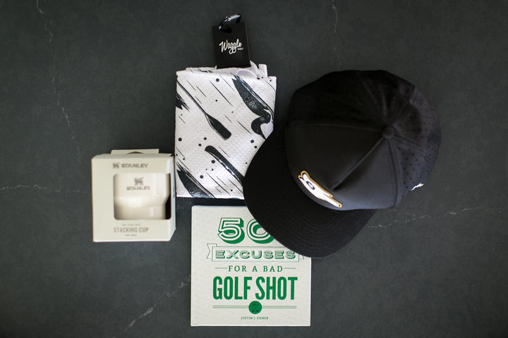 A curated flat-lay of golf-themed merchandise on a dark slate background, featuring a black baseball cap with a gold logo, a white and black patterned towel, a boxed golf cup by Stanley, and a green and white golf excuse card.