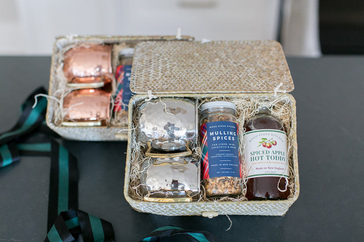 Gift basket with apple hot toddy cocktail mix, choice of copper or silver mugs in a woven gift box set on a dark surface.