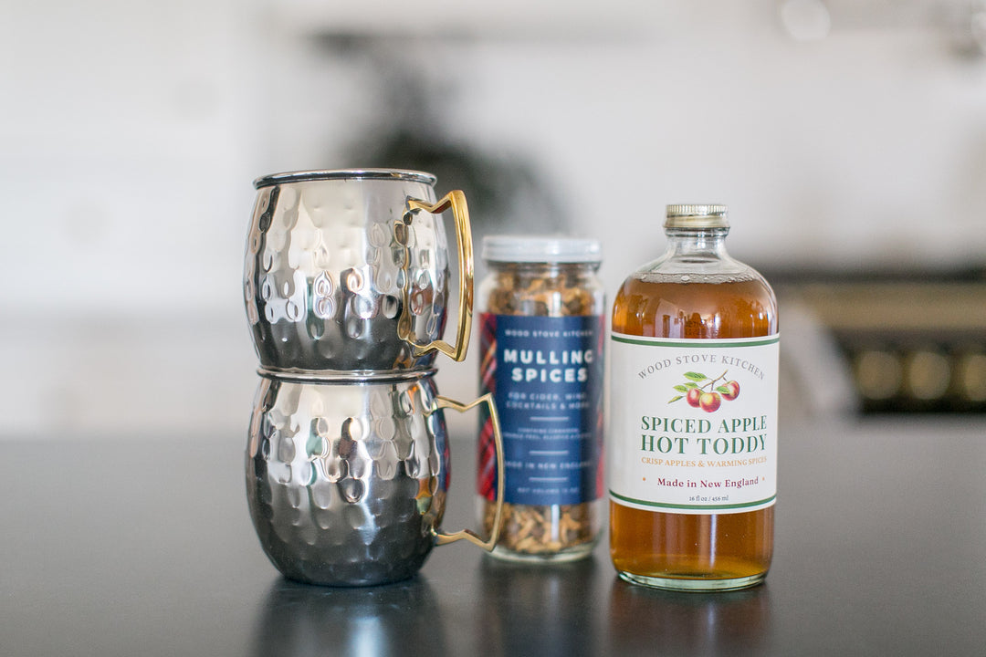 Two silver mugs, a bottle of spiced apple hot toddy, and a container of mulling spices on a surface.