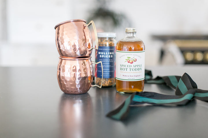 Spiced apple hot toddy bottle with copper mugs and a jar of spices on a table.
