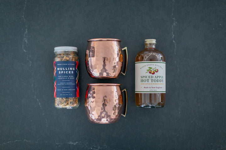 Two copper mugs and a bottle of spiced apple hot toddy with a jar of Mulling Spices on a dark slate background
