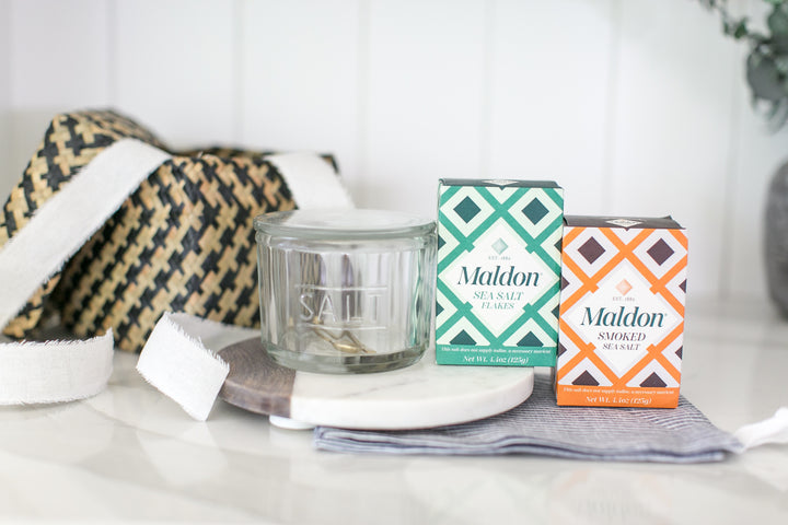 Maldon Sea Salt Flavors products displayed on a white marble tray, including a clear glass jar of sea salt, a green and white patterned box, and an orange and white patterned box, with a woven textile and white soap in the background.