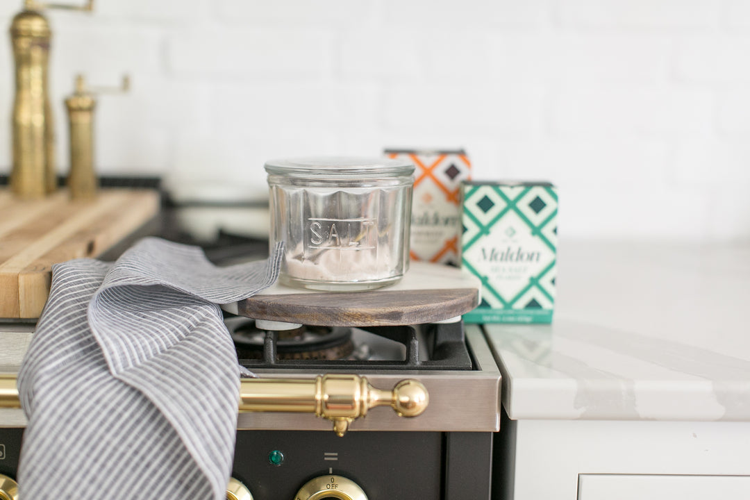 A clear glass jar candle with a gray lid sits on a small wooden coaster on top of a kitchen stove. A gray striped tea towel is draped over the front, with two boxes of Stalden tea in the background on a white countertop.