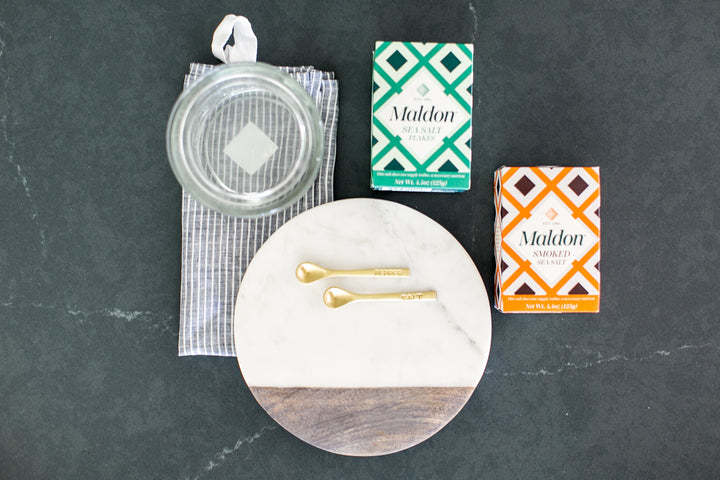 A styled flat-lay of kitchen items on a dark slate surface, including a clear glass jar lid with a white handle, a green and white patterned box of Maldon Flavored Salt, an orange and white patterned box of Maldon Asparagus Flavored Salt, a round white marble and wood serving board, and two gold spoons.