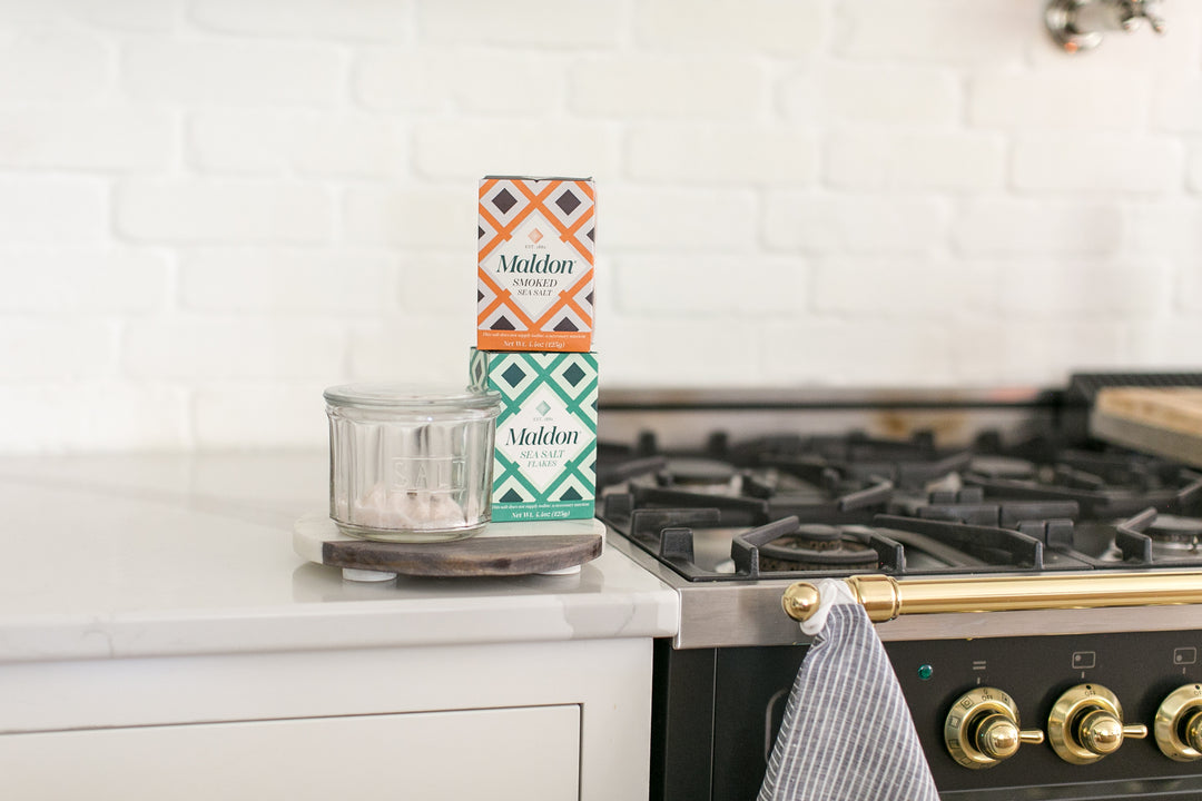 Two boxes of Maldon Sea Salt stacked vertically on a small glass jar with a wooden saucer, set on a white kitchen countertop next to a gas stove.