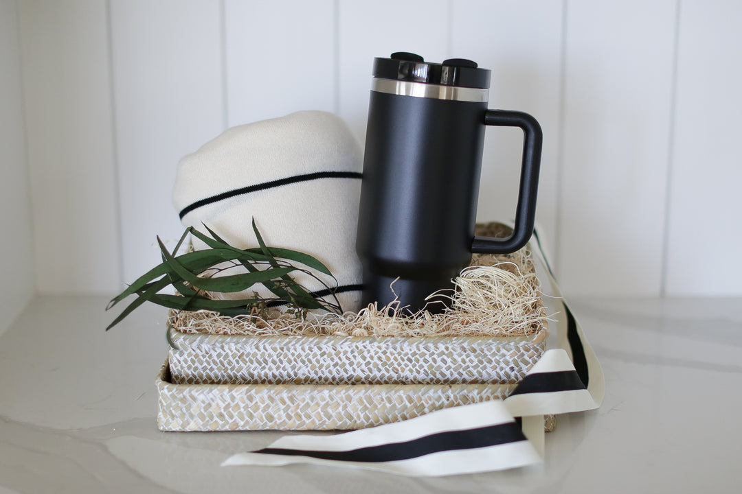 A woven gift basket containing a black and white striped knit throw blanket and a black insulated tumbler with a handle, placed on a shelf with a white background.