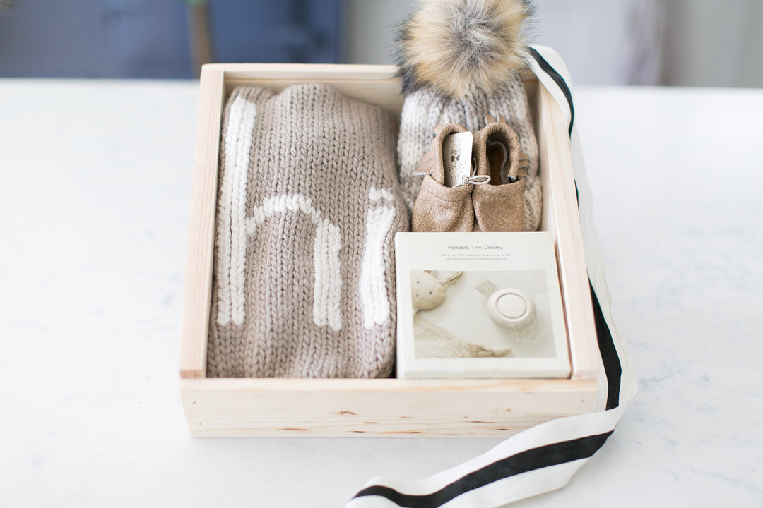 Close-up view of a Signe Gift Box, a customizable pine box with lid and a black bow, overflowing with newborn essentials: a knit blanket, a plaid hat, a sound machine, moccasins, and a keepsake box