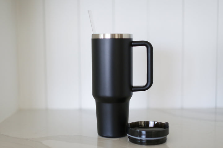A black 40oz tumbler with straw, handle, and a lid thats on the ground. Placed on a marble countertop and a shiplap background.
