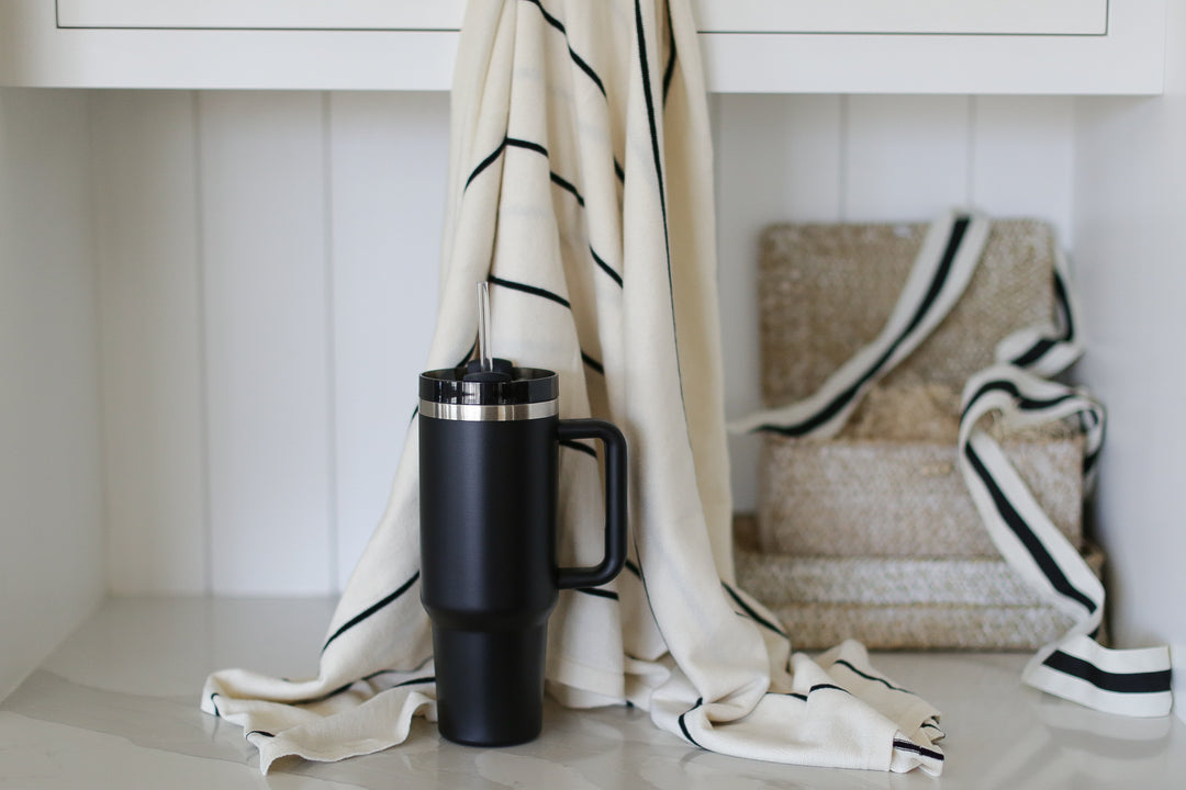 A striped cream and black blanket hung from a white cabinet. A black tumbler in front of the blanket. white walls, ribbon, and gift boxes in the background.