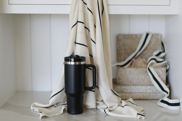 A striped cream and black blanket hung from a white cabinet. A black tumbler in front of the blanket. white walls, ribbon, and gift boxes in the background.