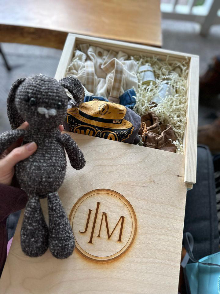 A wooden box with clothes, shoes and other oils inside. A dark gray bunny stuffed animal being held next to the box.