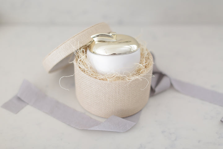 Brass painted ceramic candle in a round woven paper box with gray ribbon. 