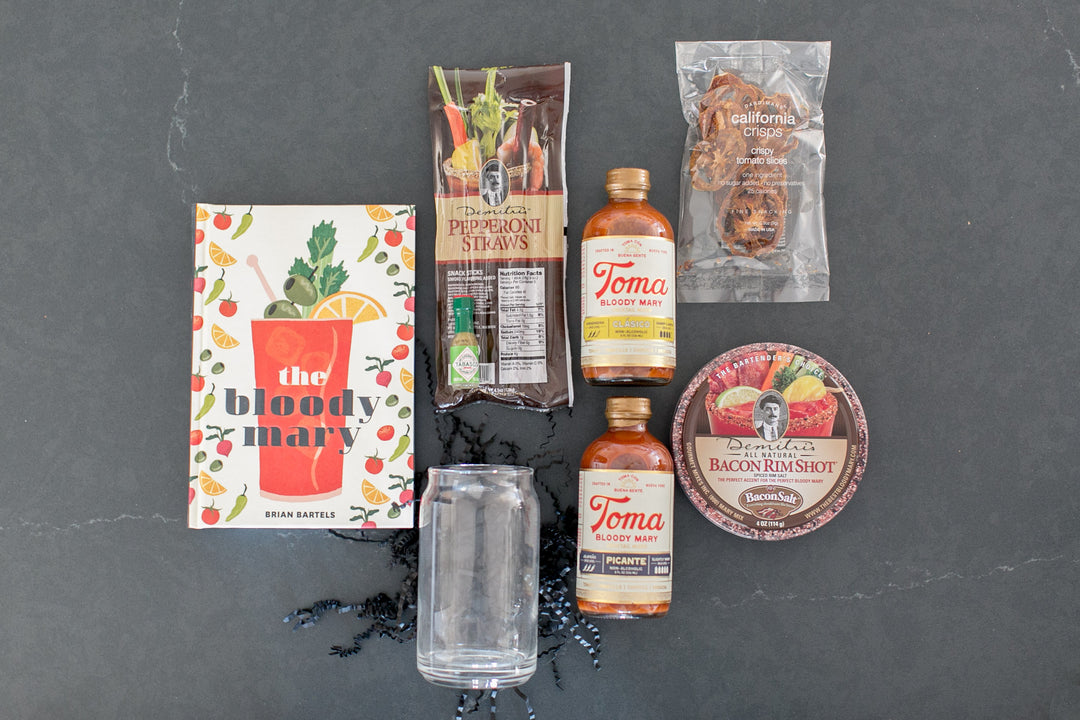 Assorted food items and a cocktail book on a dark surface. Toma brand bloody mary mix with bacon rimshot dried tomatoes, pepperoni straws and bloody mary book