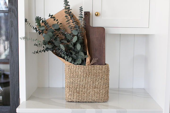 Woven basket with kitchen items including a walnut stained mango wood board that can be personalized and a preserved eucalyptus bouquet.