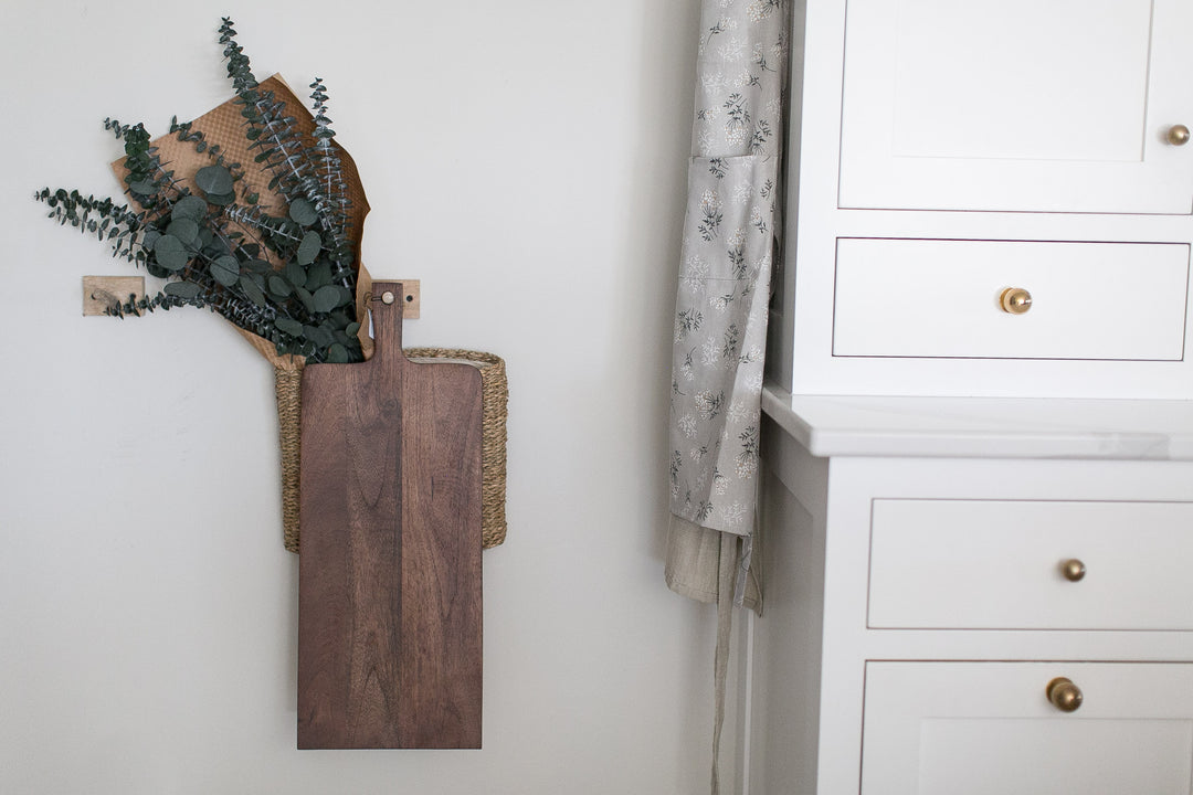 Wooden cutting board with preserved eucalyptus on a white wall next to a white shaker style inset cabinets with unlacquered brass knobs.