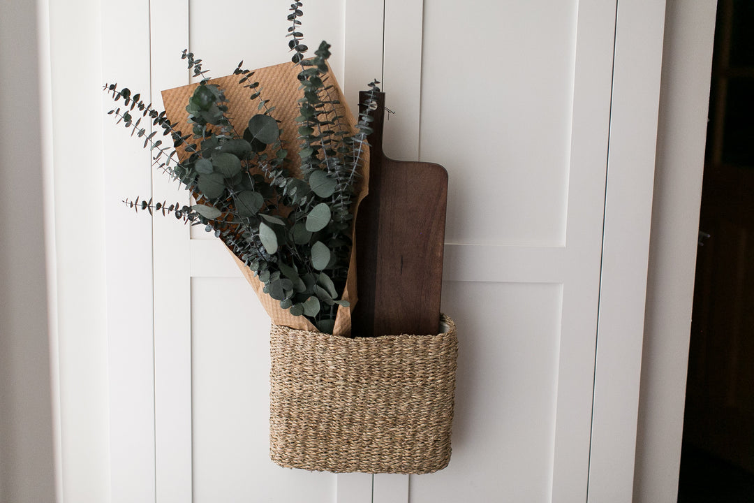 Woven basket with preserved eucalyptus greenery and a wooden cutting board on a white paneled cabinet.